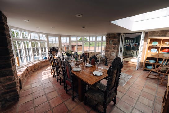 Sunlit Notehouse Farms dining room with stone walls, terracotta tiled floor, a long set wooden table, and large windows overlooking the courtyard
