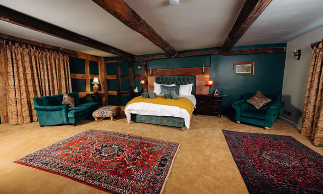 Wide-angle view of a Notehouse Farms bedroom with exposed wooden beams, dark teal walls, patterned rugs, and a bed with a mustard throw