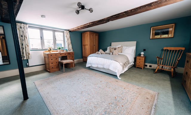 Wide-angle view of a Notehouse Farms bedroom with teal walls, an exposed wooden beam, wooden furniture, a rocking chair, and a large pale rug