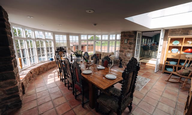 Sunlit Notehouse Farms dining room with stone walls, terracotta tiled floor, a long set wooden table, and large windows overlooking the courtyard